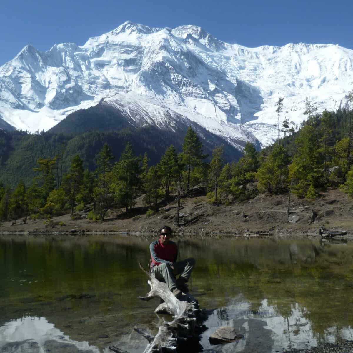 Hiker sitting on a tree trunk by a mountain lake with snowy peaks in Annapurna region, Nepal