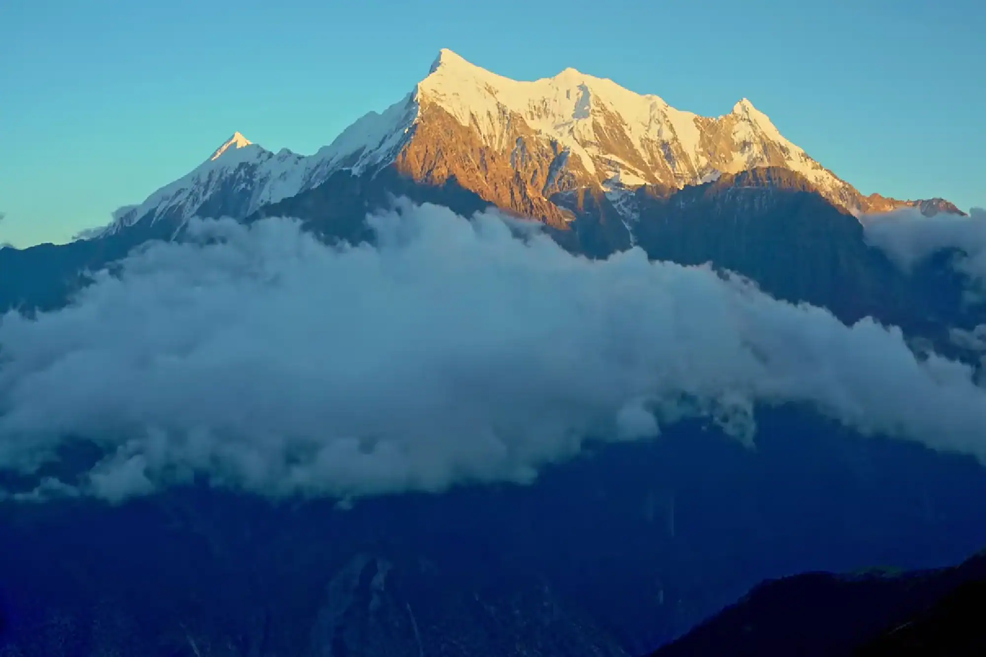 Snow-capped mountain peak glowing in golden sunrise light above thick white clouds
