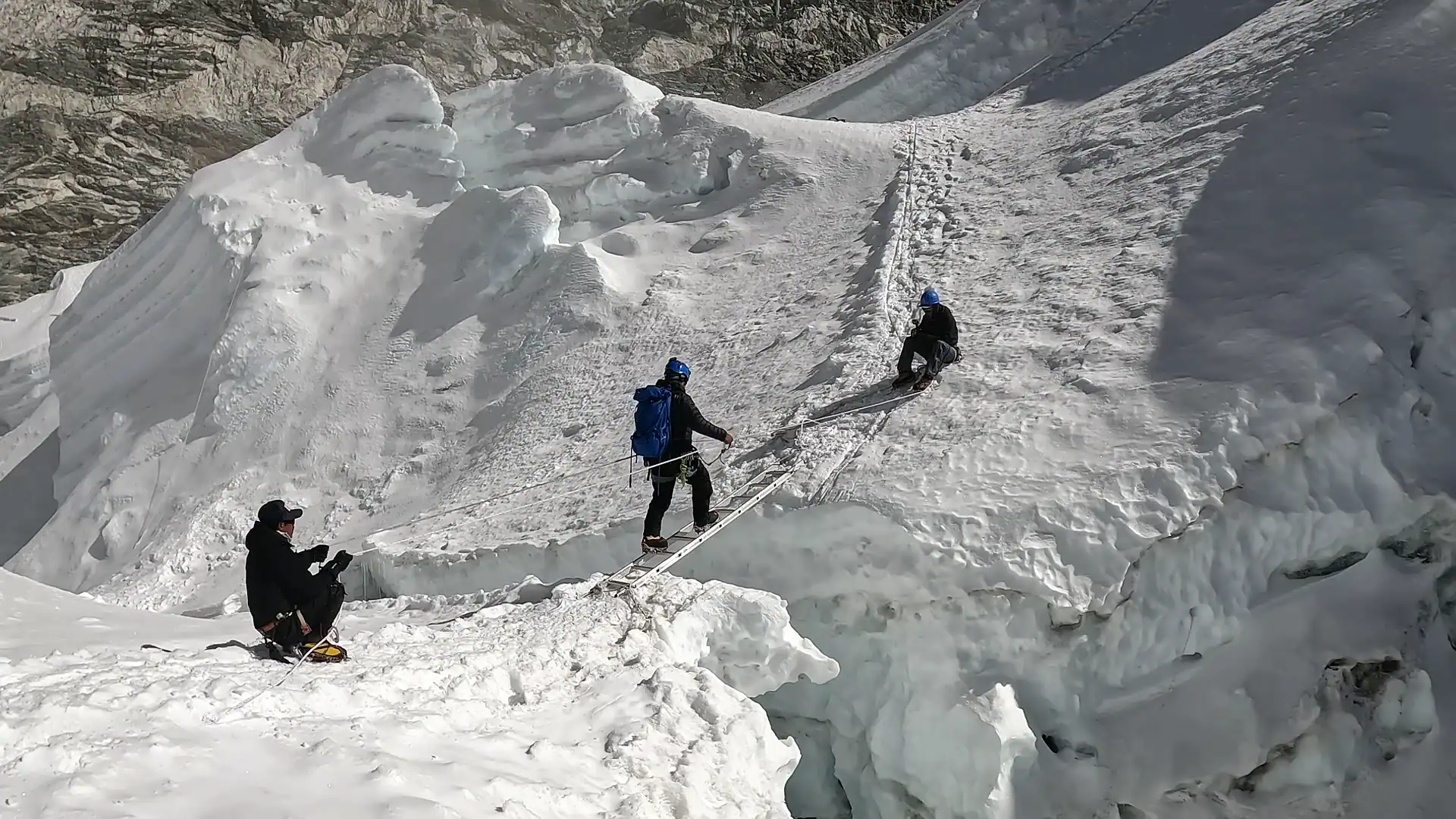 Mountaineer crossing a deep glacier crevasse on a ladder bridge along a snowy Himalayan climbing route.