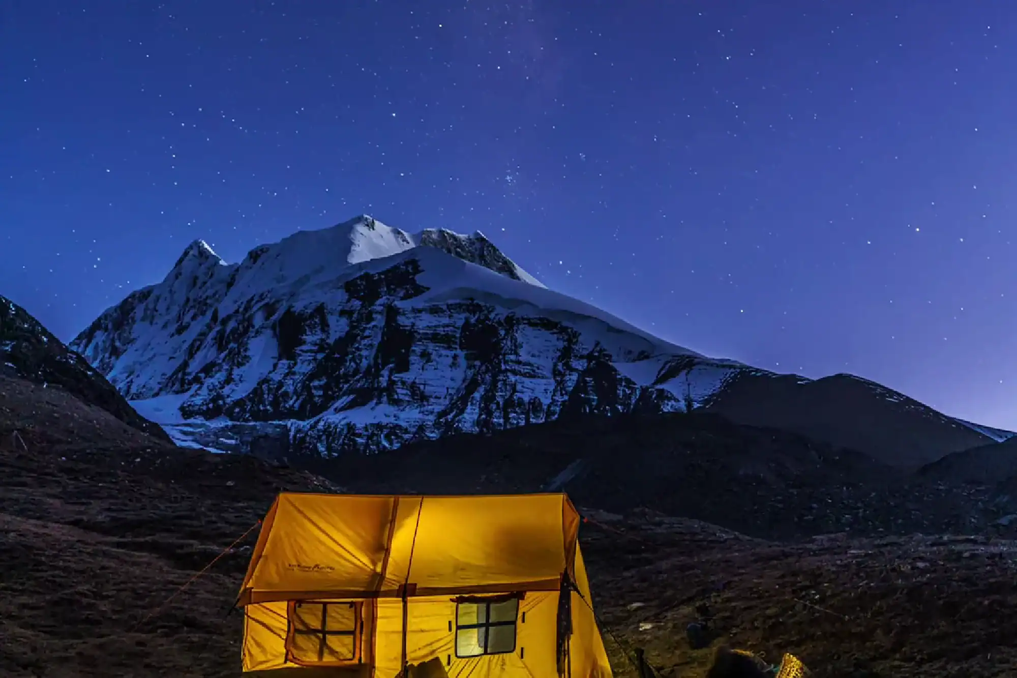 Illuminated yellow camping tent under a starry night sky with snow-covered mountain peaks in the background.