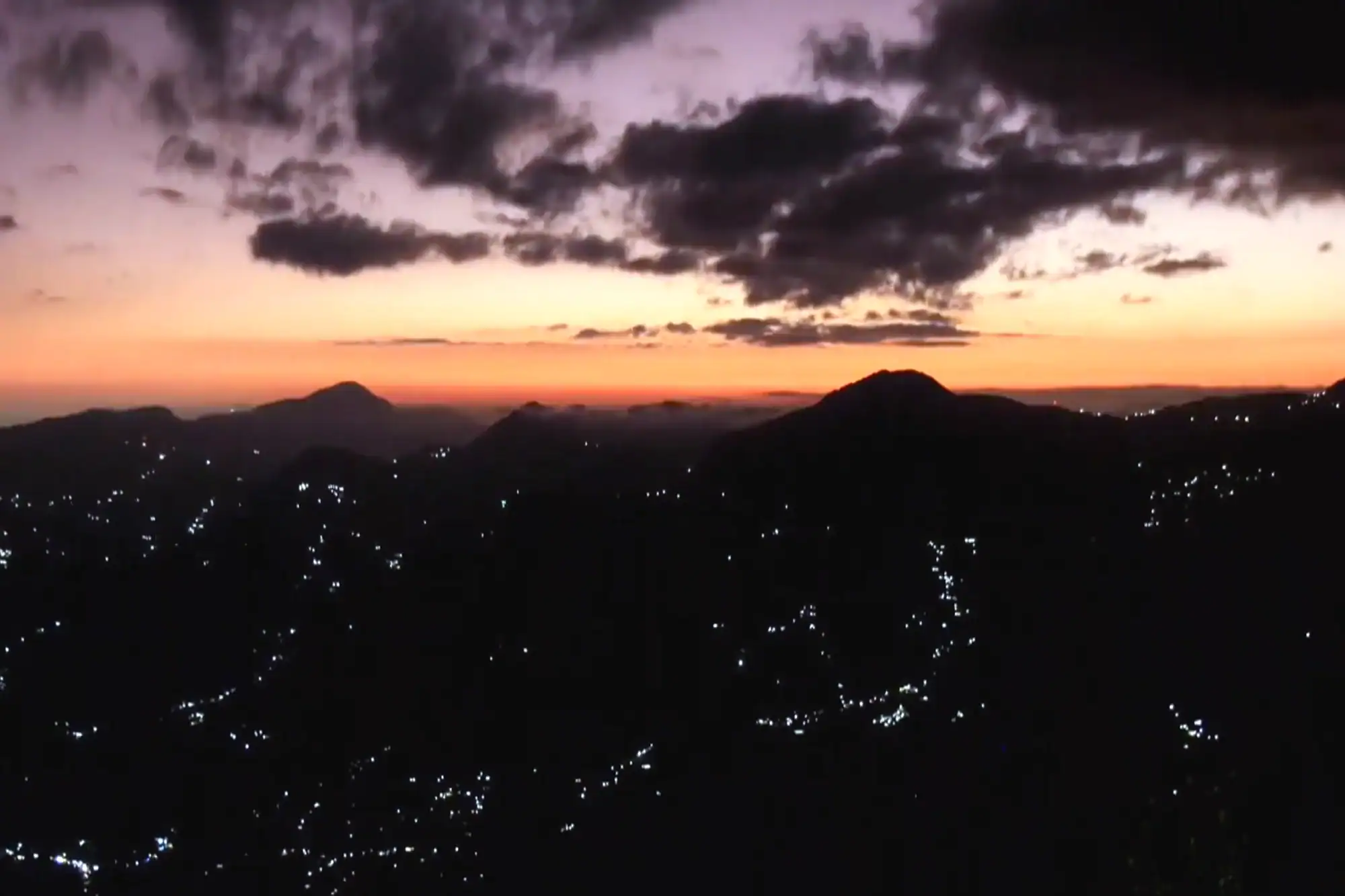 Silhouetted mountains with glowing night lights beneath a dramatic orange and purple twilight sky.