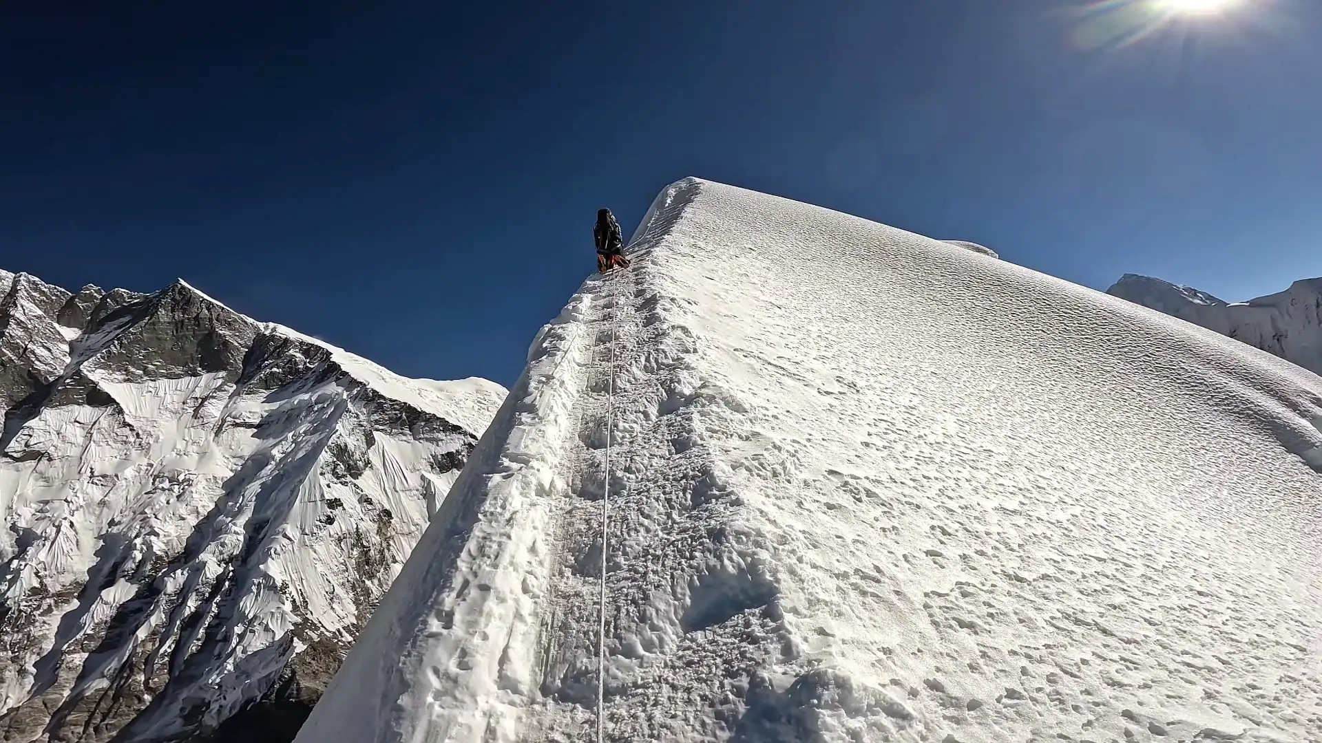 Climber ascending a steep snowy mountain ridge with fixed ropes under a clear blue sky