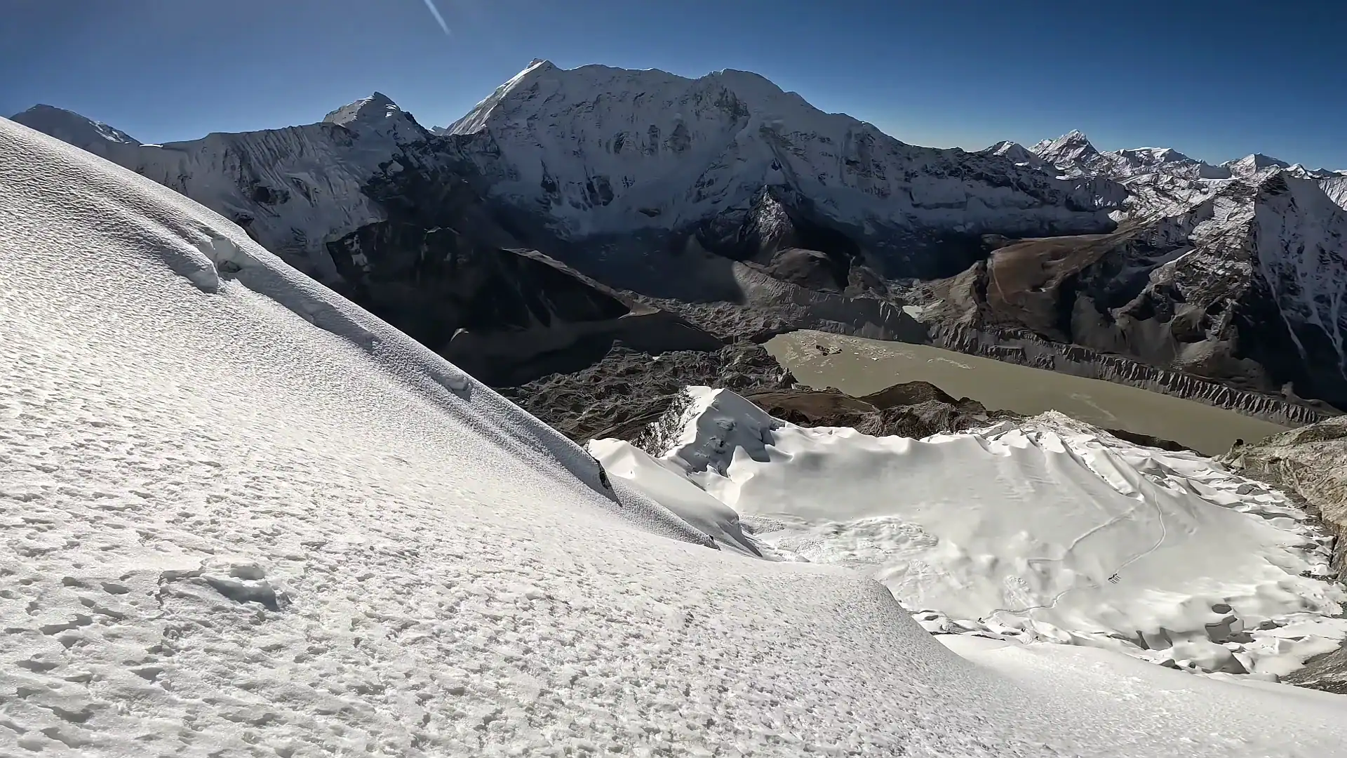 Snow-covered mountain ridge overlooking a glacial lake and rugged high-altitude peaks under a clear blue sky.