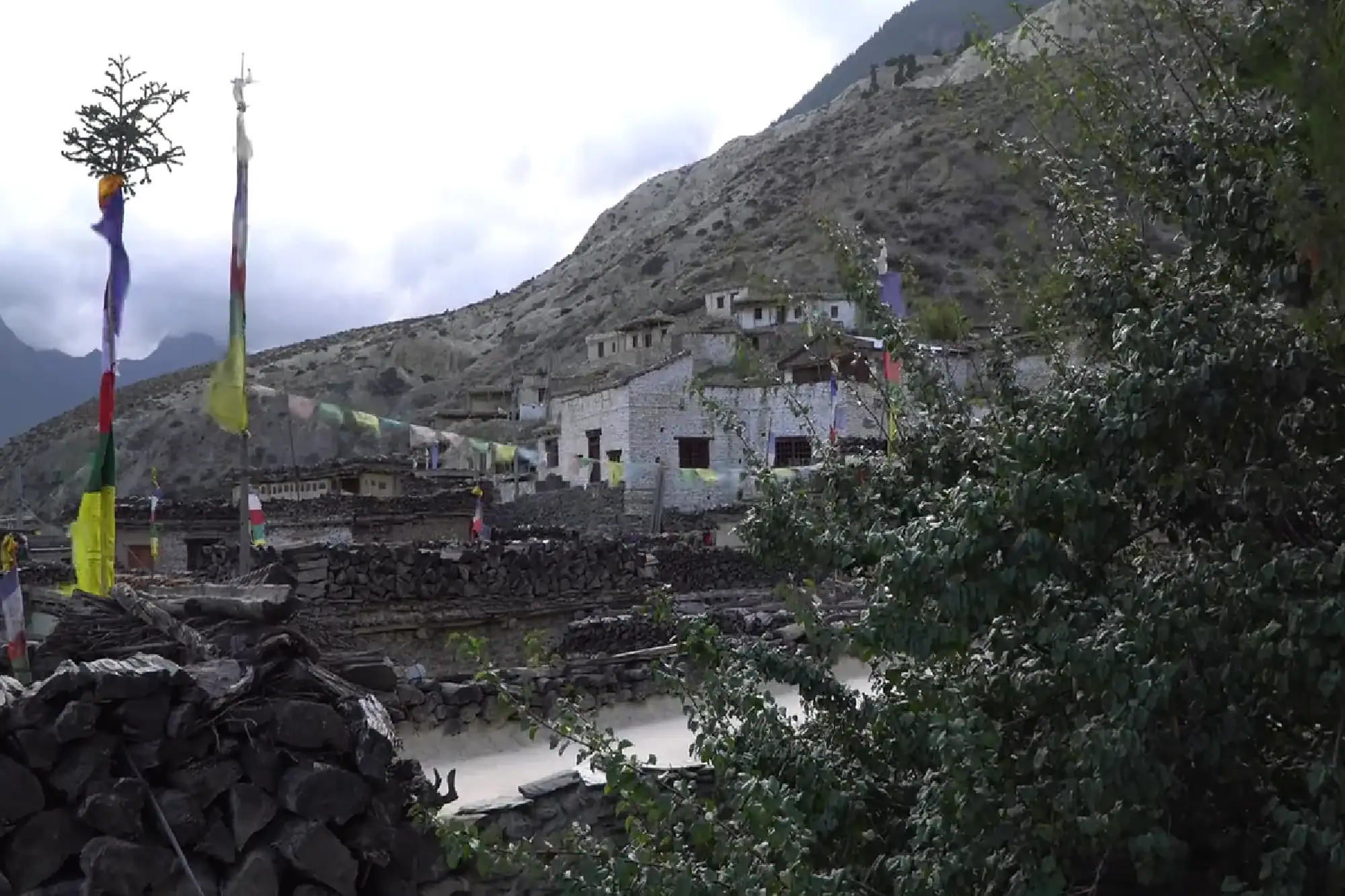 Traditional stone houses and colorful Buddhist prayer flags on a rugged mountain hillside in Nepal.