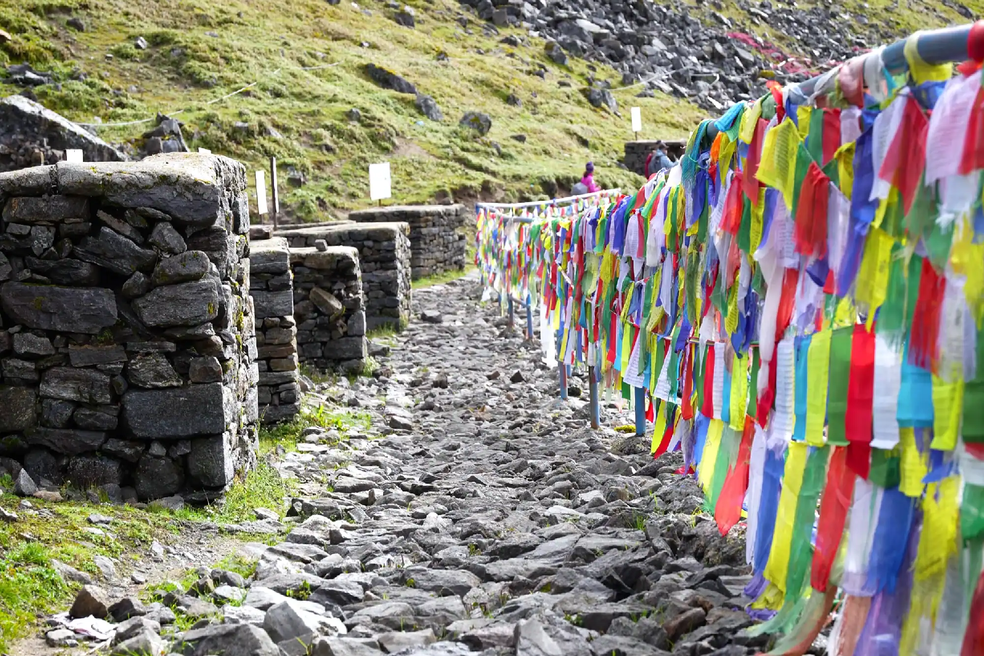 Colorful Buddhist prayer flags along a rocky Himalayan mountain trail with stone wall borders.