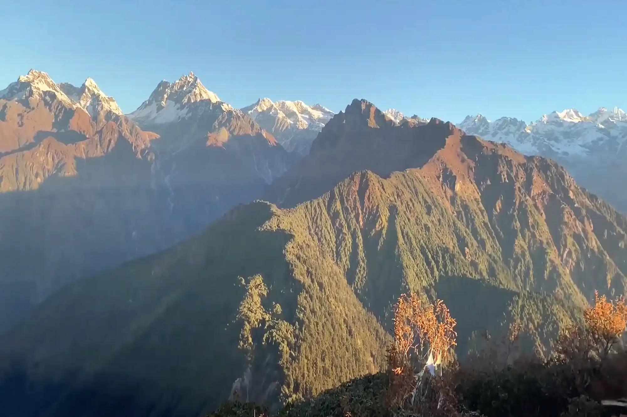 Panoramic view of rugged Himalayan mountain ranges with snow-capped peaks under a clear blue sky during sunrise.