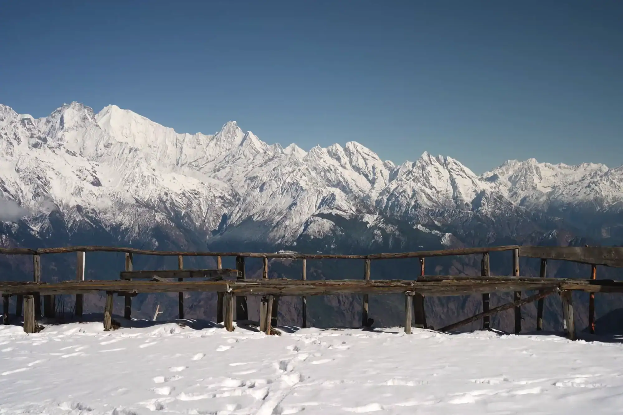 Rustic wooden bench on a snowy mountain viewpoint overlooking majestic snow-covered Himalayan peaks against a clear blue sky.