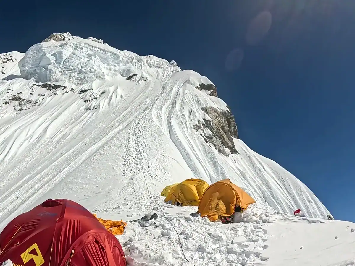 Mountaineering expedition camp with yellow and red tents on a snowy ridge below a steep mountain peak.