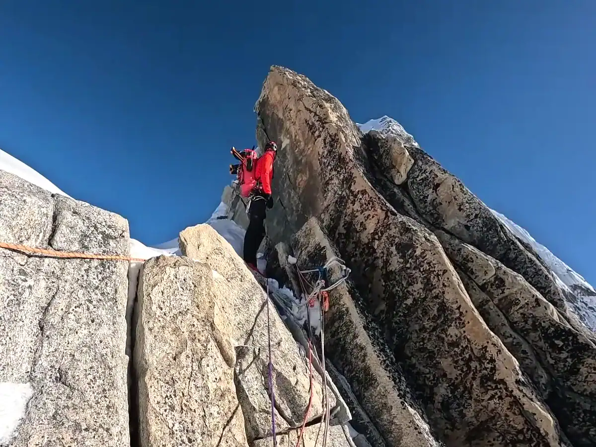 Climber ascending a steep rocky ridge with ropes on a snowy mountain peak under a clear blue sky