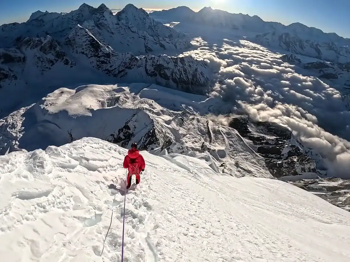 Climber in red descending a snowy mountain peak overlooking a vast alpine range above the clouds.