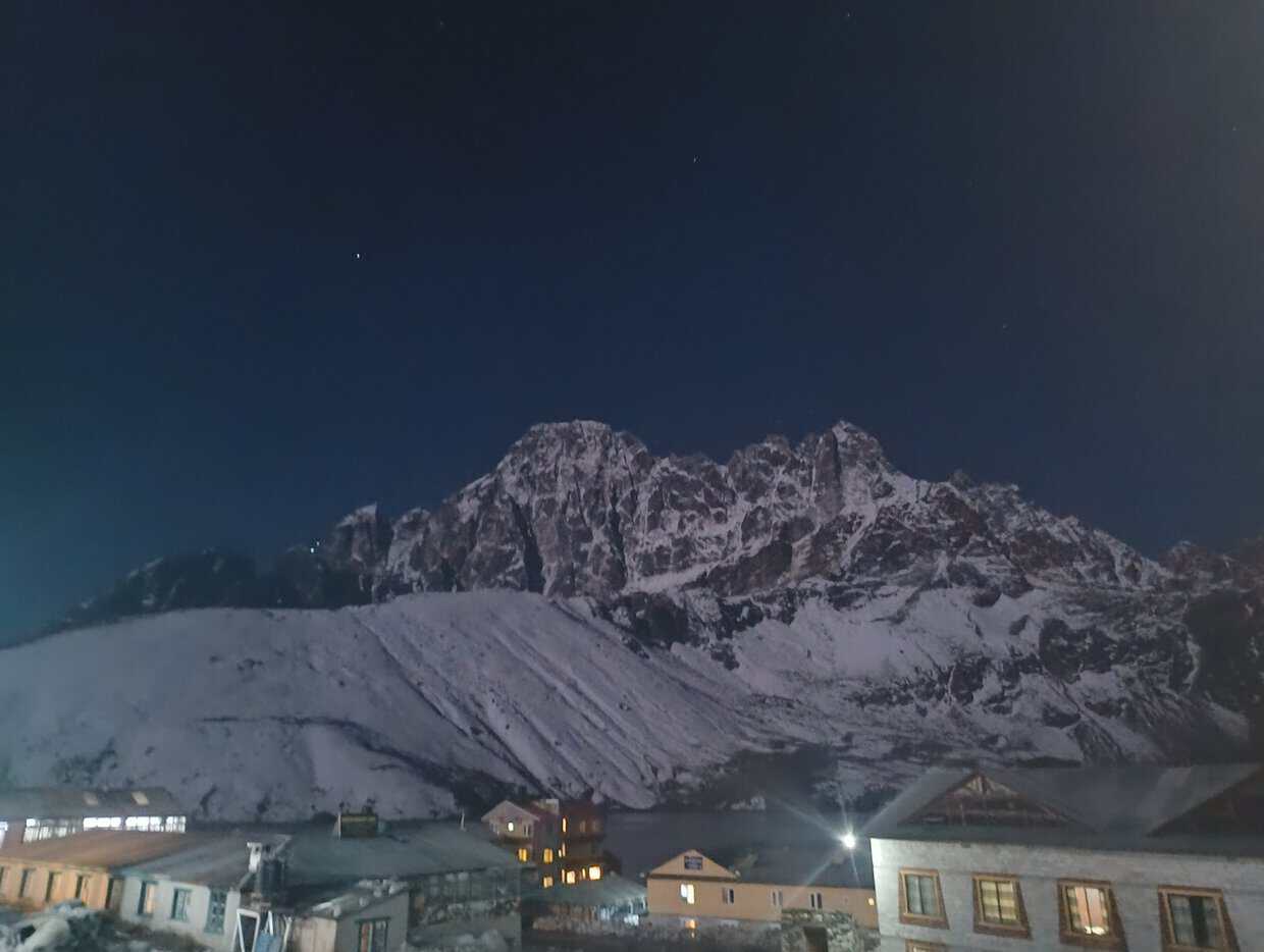 Snowy Himalayan mountain peaks towering over a illuminated mountain village at night under a clear dark sky.